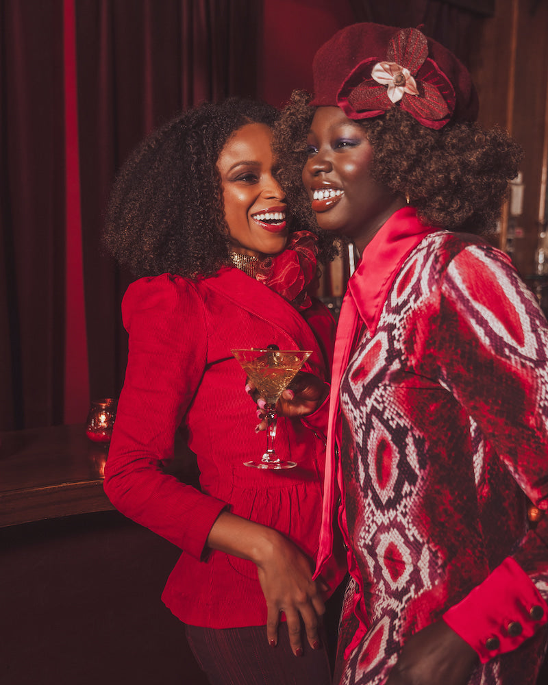 Two women in red and patterned outfits posing together with a drink in a dimly lit setting.