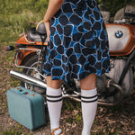 Person wearing a blue patterned dress standing next to a vintage motorcycle with a suitcase nearby wearing Soul Sister since 1969..