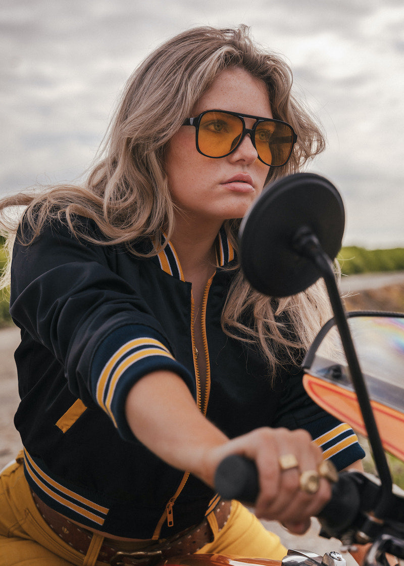 Woman wearing sunglasses and a black and yellow jacket on a motorcycle wearing Soul Sister since 1969.