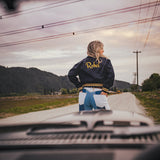 Person wearing a jacket with 'Rebel' on it standing on a road with mountains in the background wearing Soul Sister since 1969