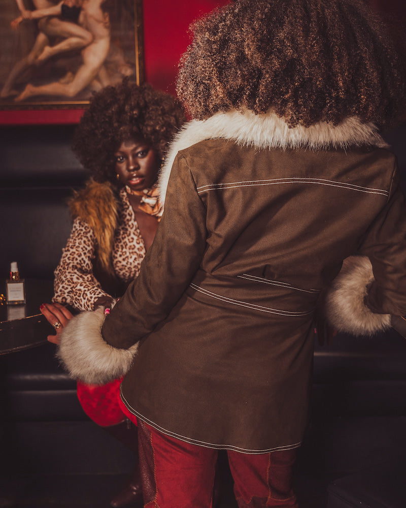 Person wearing a brown coat with fur trim interacting with another person in a dimly lit room.