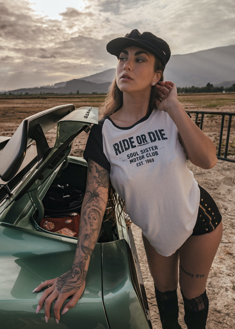 Woman wearing a 'Ride or Die' t-shirt leaning against a car in a desert setting wears clothing by Soul Sister since 1969