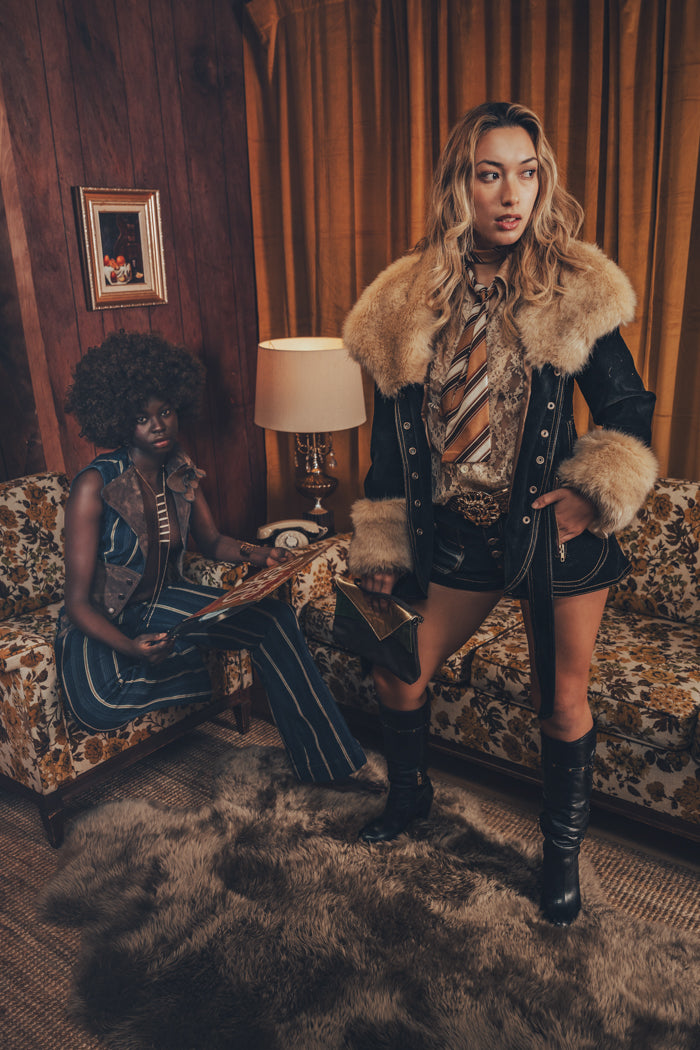 A person wearing a faux fur lined coat with gold satin detailing and bold topstitching, paired with a patterned shirt and shorts.