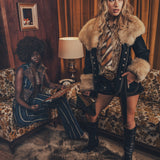 A person wearing a faux fur lined coat with gold satin detailing and bold topstitching, paired with a patterned shirt and shorts.