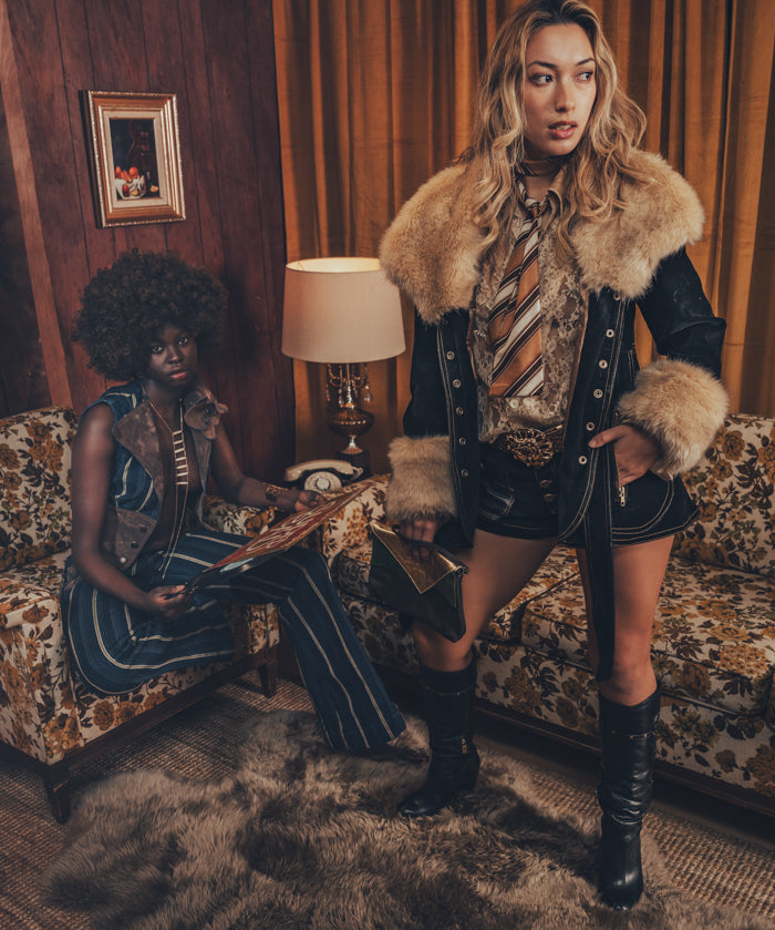 A person wearing a faux fur lined coat with gold satin detailing and bold topstitching, paired with a patterned shirt and shorts.