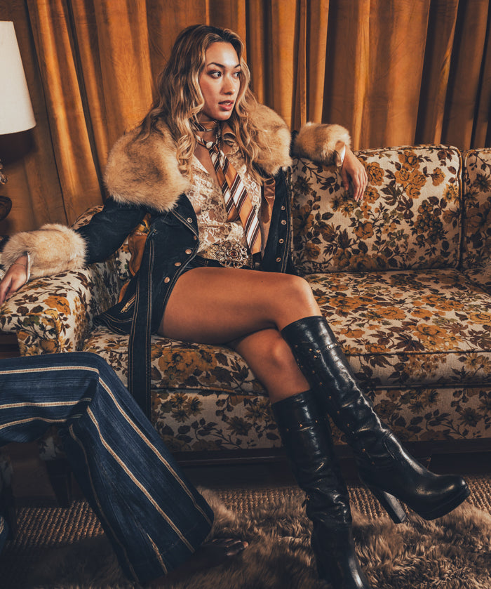 A person wearing a faux fur lined coat with gold satin detailing and bold topstitching, paired with a patterned shirt and shorts.