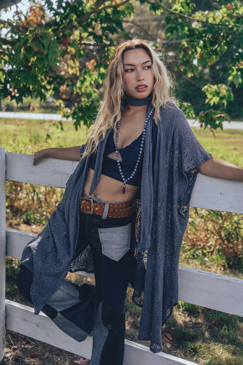 A woman wearing a knit crochet lace robe in gray, with a skinny scarf, paired with jeans and standing by a fence in a natural outdoor setting.