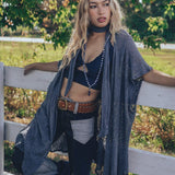 A woman wearing a knit crochet lace robe in gray, with a skinny scarf, paired with jeans and standing by a fence in a natural outdoor setting.