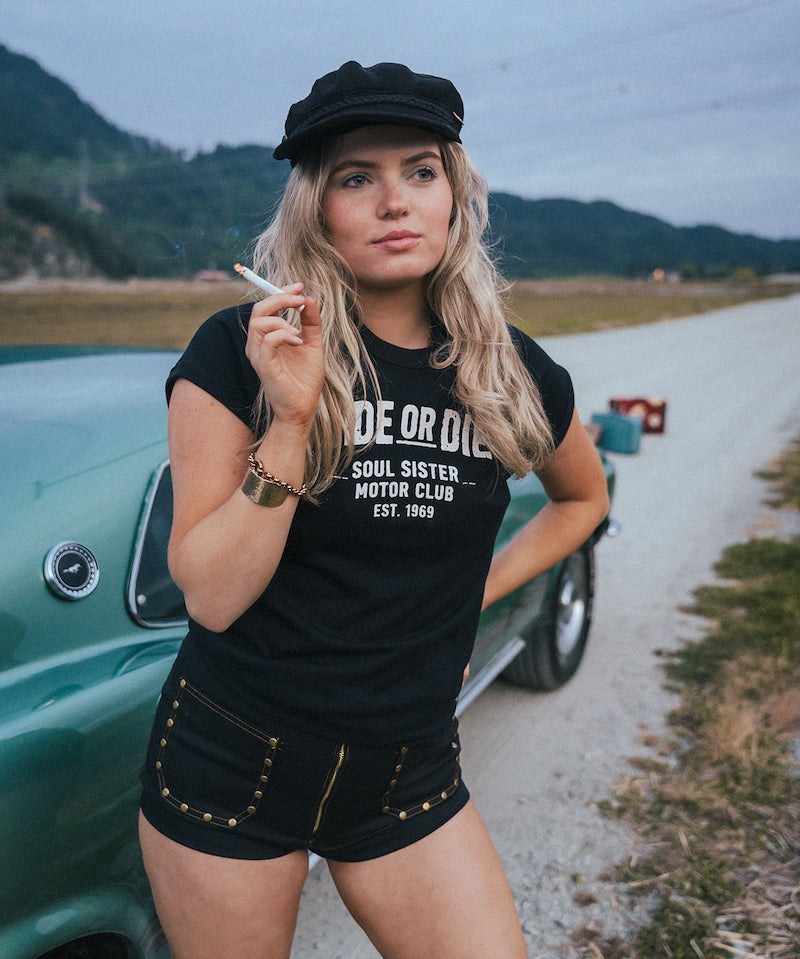 Woman in black cap and t-shirt standing next to a vintage car on a road with mountains in the background Ride or Die Motor Club wearing soul sister since 1969
