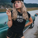Woman in a black t-shirt with text leaning against a green car outdoors Ride or Die Motor Club wearing soul sister since 1969.