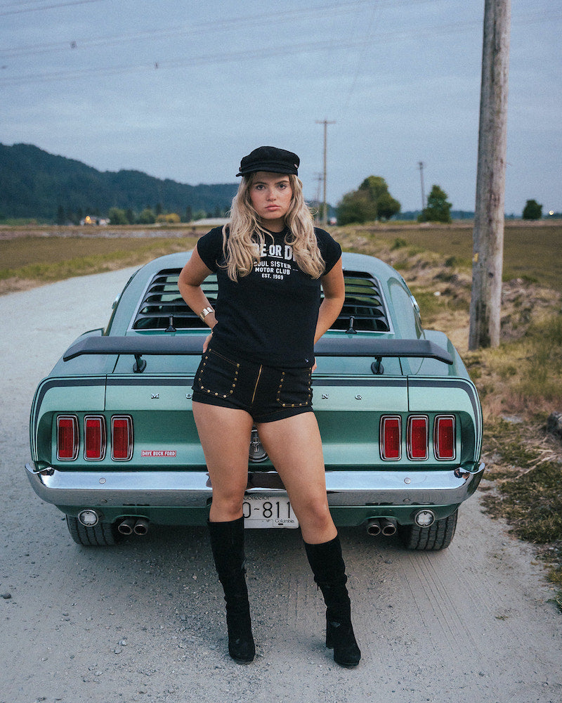 Woman posing in front of a vintage car on a rural road Ride or Die Motor Club wearing soul sister since 1969