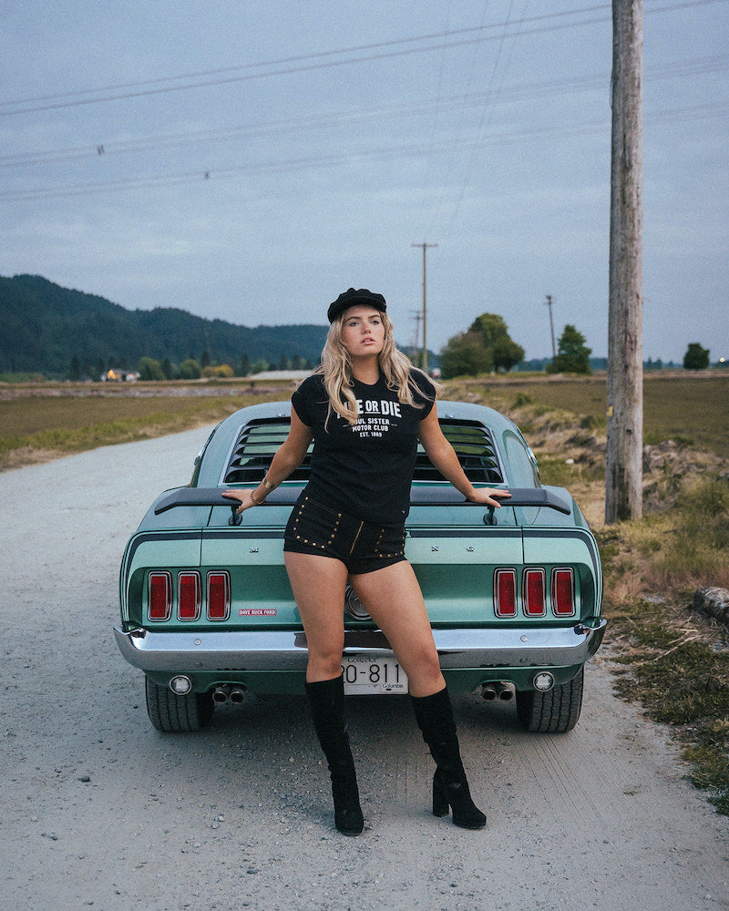 Woman standing next to a classic car on a rural road Ride or Die Motor Club wearing soul sister since 1969