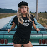 Woman wearing a black 'De or Die' t-shirt standing next to a vintage car on a road Ride or Die Motor Club wearing soul sister since 1969.