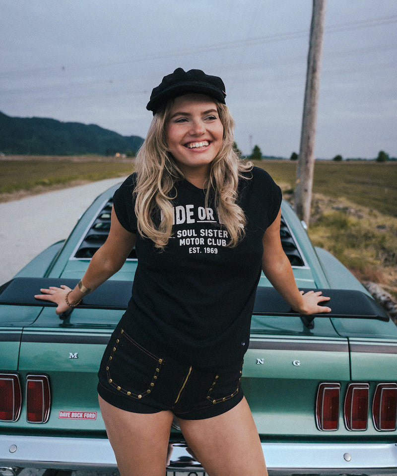 Woman standing next to a vintage car wearing a black t-shirt with text Ride or Die Motor Club wearing soul sister since 1969