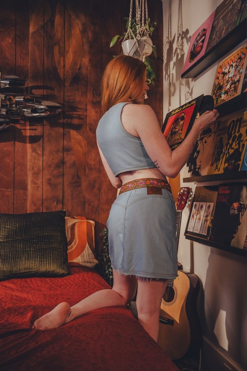 Woman in a casual outfit standing in a room with a guitar and shelves.