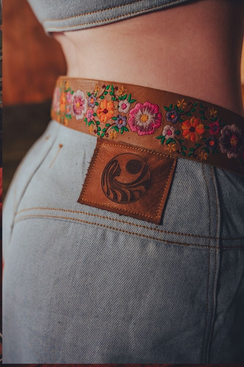 Close-up of a person wearing a floral embroidered belt with a leather patch on jeans.