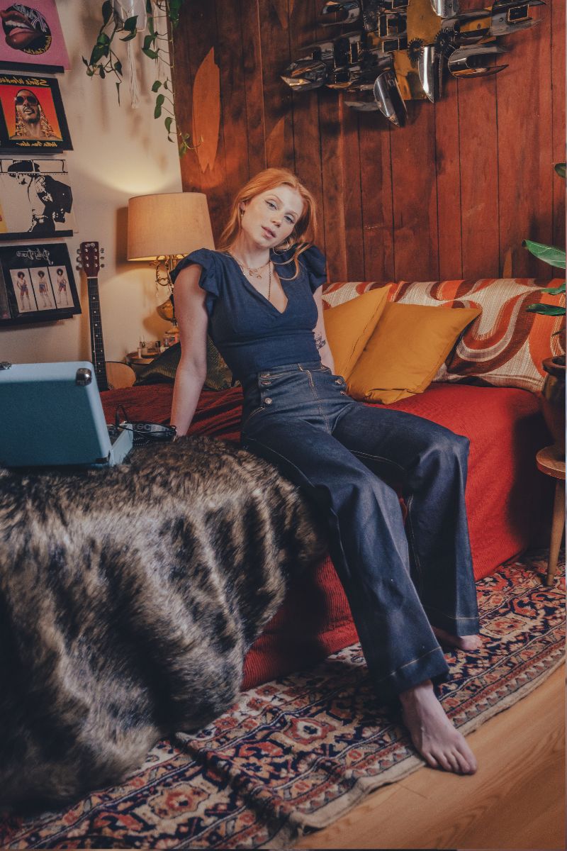 Woman sitting on a 1970s style couch in a cozy living room with a vintage record player wearing high rise sailor bell bottom jeans.