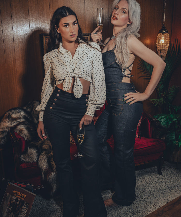 2 chic women in coordinated black 70's ensemble poses in front of a wood panelled wall and mid century furniture.