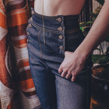 Person wearing a beige bralette and high-waisted blue jeans against a patterned curtain background.