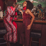 Two women in a bar setting, one in a patterned red dress and the other in a solid red dress, enjoying drinks.