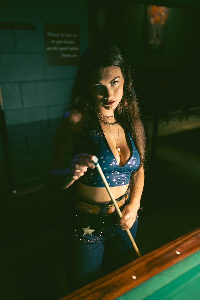 Woman holding a pool cue in a dimly lit room wearing sexy high waisted bell bottom jeans