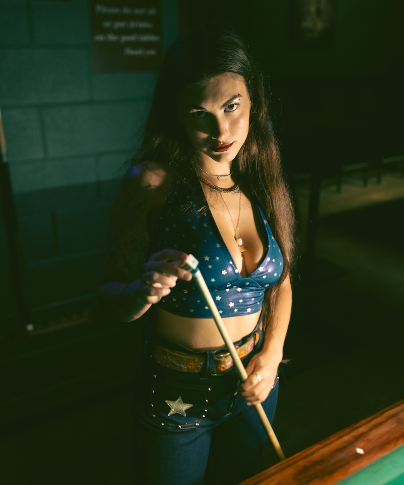 Woman holding a pool cue in a dimly lit room wearing sexy high waisted bell bottom jeans