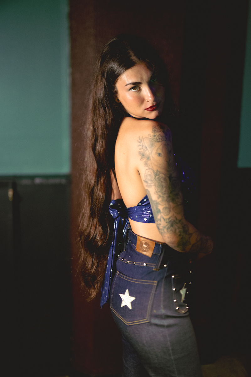 Woman wearing a 1970s inspired halter top with stars a blue top and denim bellbottom jeans with star details, standing against a dark retro inspired background.