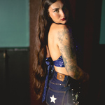 Woman wearing a 1970s inspired halter top with stars a blue top and denim bellbottom jeans with star details, standing against a dark retro inspired background.