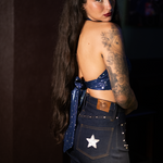 Woman wearing a blue 1970s halter top  and dark  bellbottom jeans with a star patch, standing against a dark background.