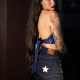 Woman wearing a blue 1970s halter top  and dark  bellbottom jeans with a star patch, standing against a dark background.