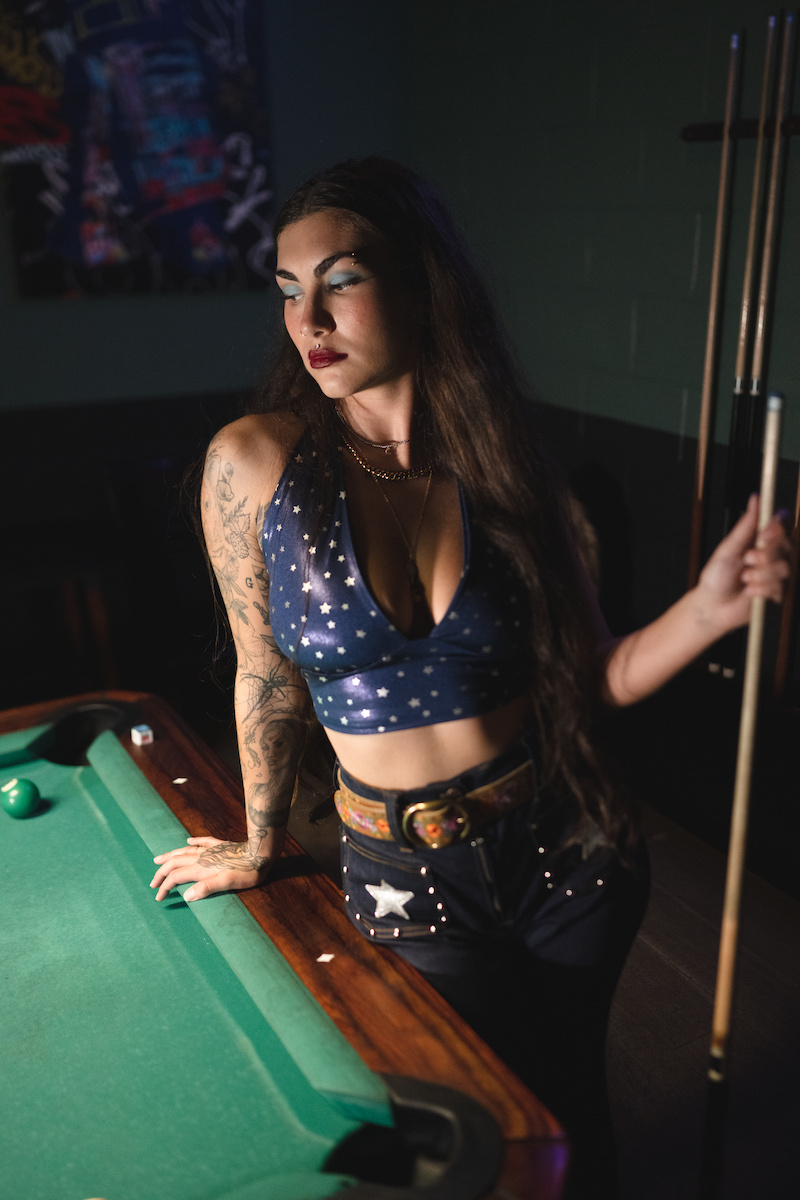 Woman in a retro blue star-patterned top  and funky high waisted studded bell bottom jeans, standing next to a pool table holding a cue.