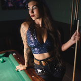 Woman in a retro blue star-patterned top  and funky high waisted studded bell bottom jeans, standing next to a pool table holding a cue.