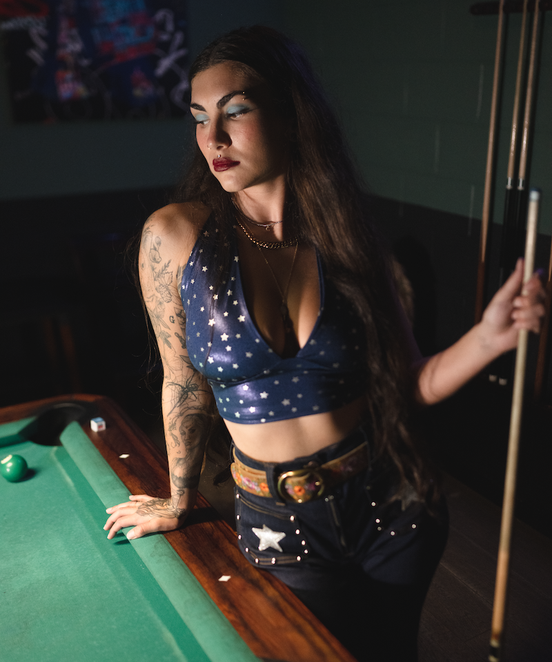 Woman in a retro blue star-patterned top  and funky high waisted studded bell bottom jeans, standing next to a pool table holding a cue.