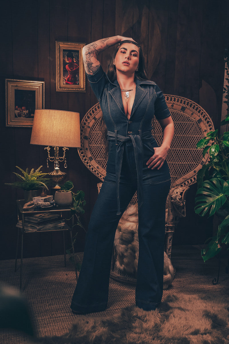 Model wearing a 70s inspired stretch denim jumpsuit with flared legs front zip closure and a plunging neckline styled for a bold retro look