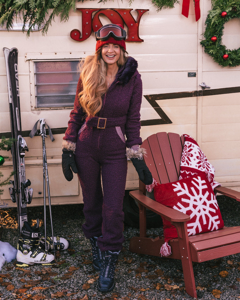 Woman in a purple snowsuit standing next to a wooden chair with a red and white blanket, in front of a trailer with Christmas decorations.