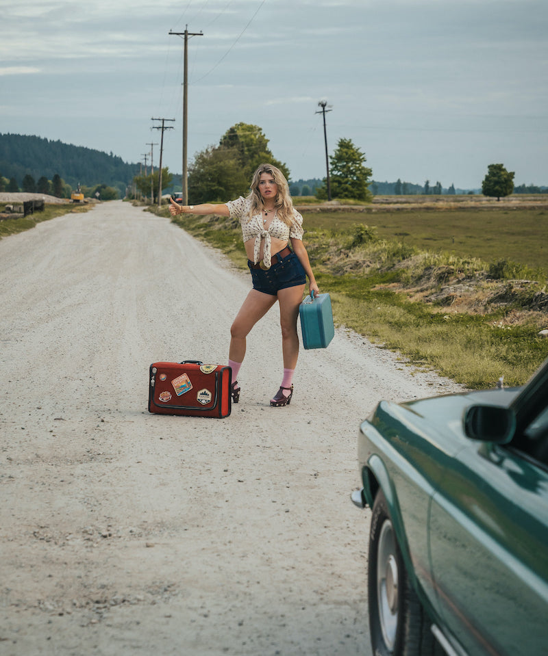 Woman standing on a dirt road with suitcases, hitchhiking wearing Soul Sister since 1969..
