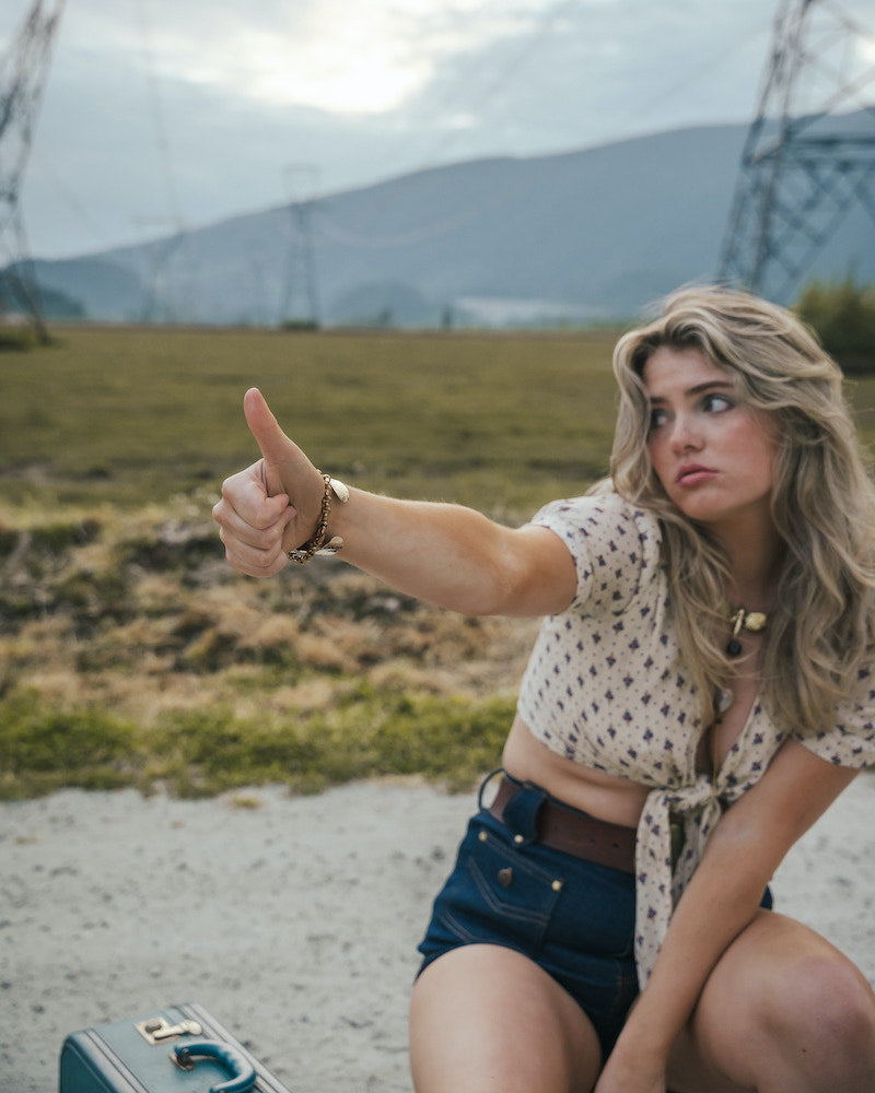 Woman sitting on a gravel road with mountains in the background, giving a thumbs-up wearing Soul Sister since 1969.