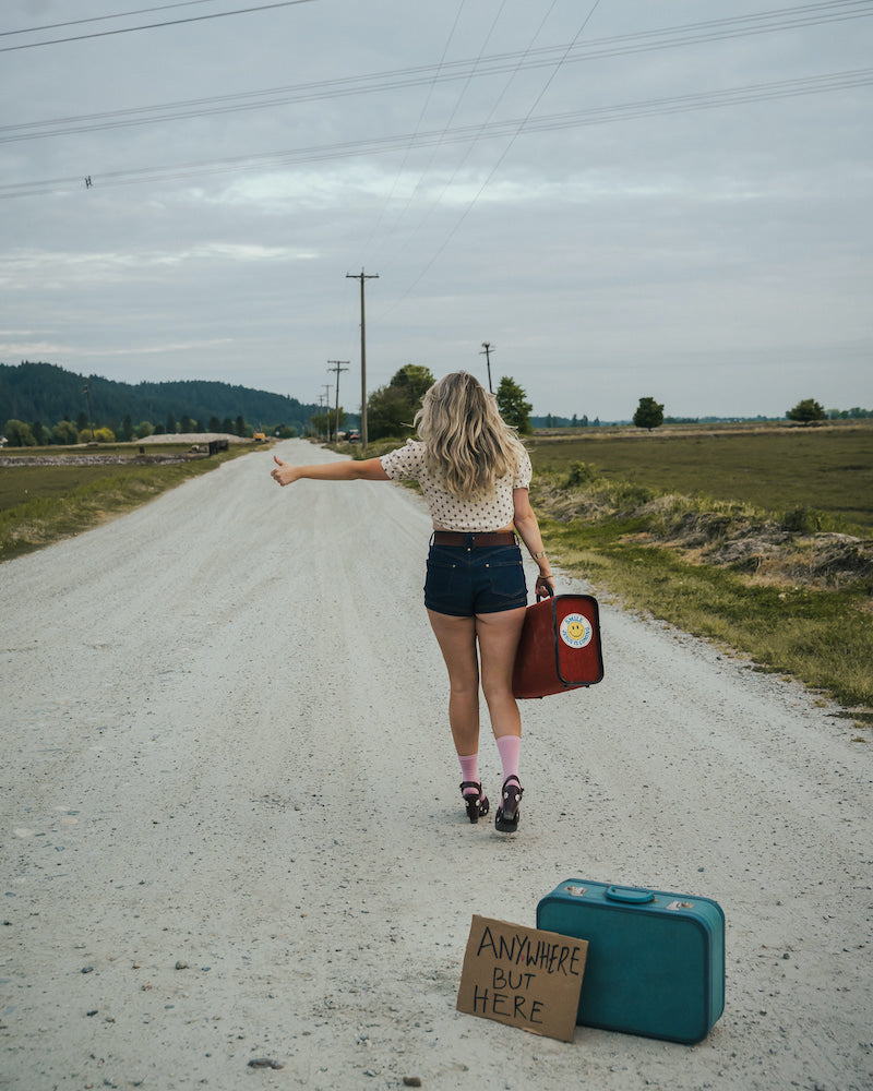 Woman standing on a road with suitcases and a sign, indicating a journey or travel theme wearing Soul Sister since 1969..