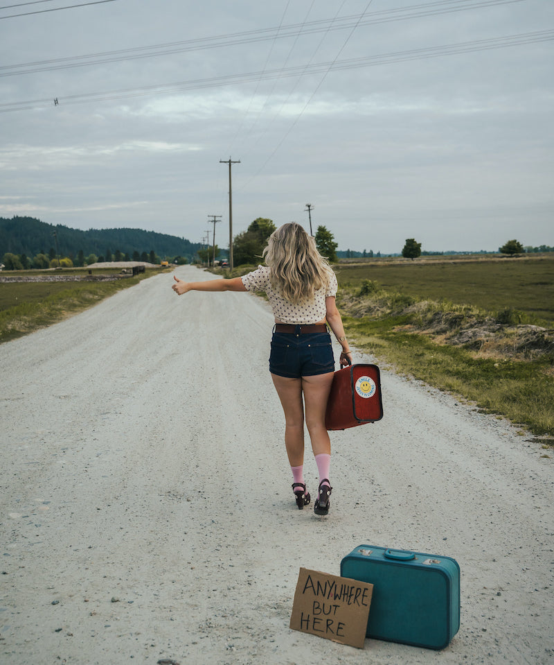 Woman standing on a road with suitcases and a sign, indicating a journey or travel theme wearing Soul Sister since 1969..