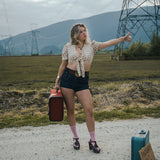 Woman hitchhiking on a road with suitcases and a sign in an open field wearing Soul Sister since 1969.