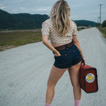 Woman standing on a road holding a red suitcase with a smiley face, wearing a polka dot shirt and denim shorts. wearing Soul Sister since 1969.