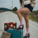 Person standing on a road with suitcases and a sign reading 'Anywhere But Here' wearing a fluffy coat and pink socks wearing Soul Sister since 1969..