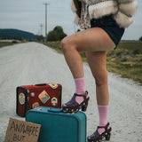 Person standing on a road with suitcases and a sign reading 'Anywhere But Here' wearing a fluffy coat and pink socks wearing Soul Sister since 1969..