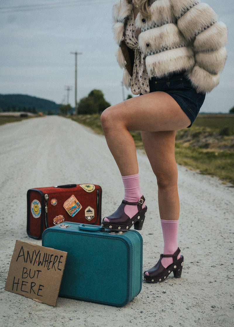 Person standing on a road with suitcases and a sign reading 'Anywhere But Here' wearing a fluffy coat and pink socks wearing soul sister since 1969.