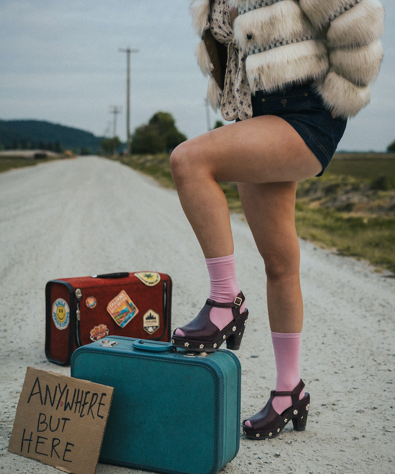 Person standing on a road with suitcases and a sign reading 'Anywhere But Here' wearing a fluffy coat and pink socks wearing soul sister since 1969.