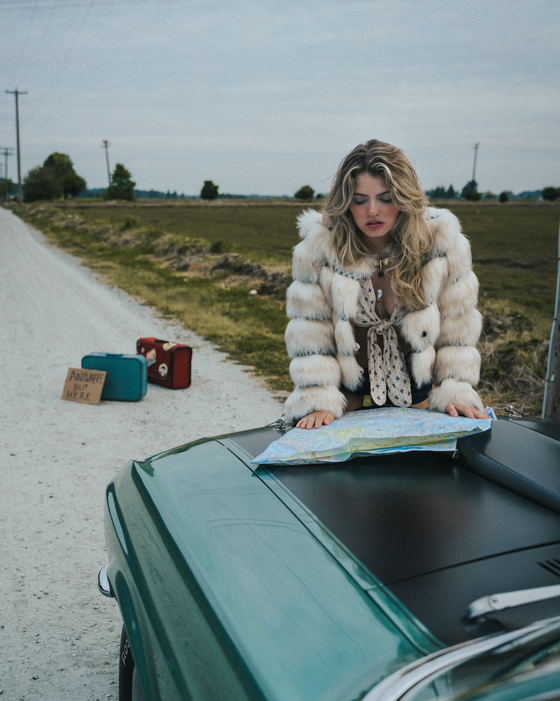 Woman in a fur coat looking at a map on a car hood by a road wearing soul sister since 1969
