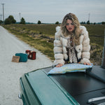 Woman in a fur coat looking at a map on a car hood by a road wearing soul sister since 1969