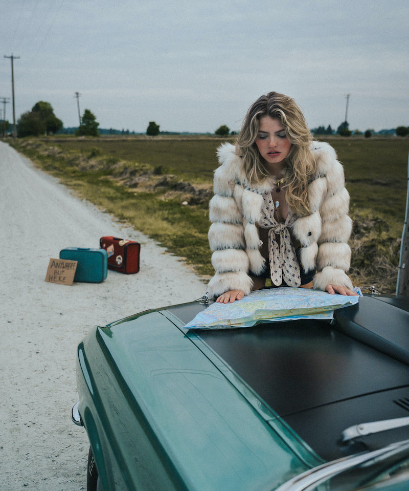 Woman in a fur coat looking at a map on a car hood by a road wearing soul sister since 1969