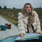 Woman in a fur coat sitting on a car hood with a map, surrounded by suitcases in an open field wearing soul sister since 1969.