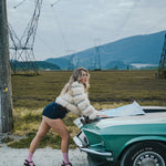 Woman in a puffer vest leaning on a vintage car in an open field with power lines and mountains in the background. wearing soul sister since 1969